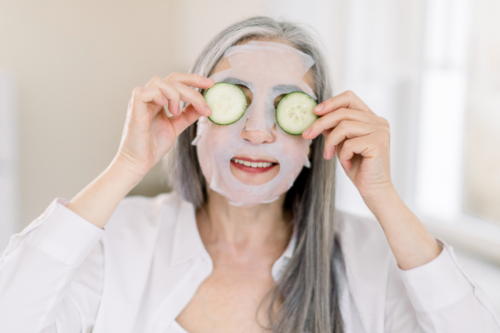a senior woman placing slices of cucumber in her eyes while wearing a face mask
